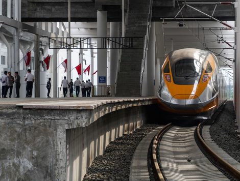A SLEEK MODERN TRAIN ENTERS ALONGSIDE A CURVING PLATFORM IN A STATION BEDECKED WITH FLAGS